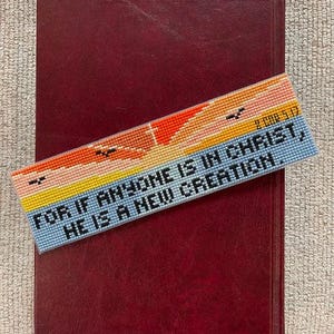 May include: A burgundy leather-bound book with a colorful, cross-stitched bookmark. The bookmark features a landscape design with text that reads, "FOR IF ANYONE IS IN CHRIST, HE IS A NEW CREATION."