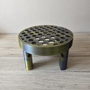 Puede incluir: Un pequeño soporte redondo para plantas con tres patas y un patrón de panal en la superficie superior. El soporte tiene un acabado metálico con tonos verdes y azules. Las patas son rectangulares e inclinadas.