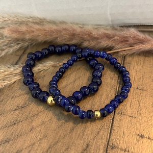 May include: Two beaded bracelets. The bracelets are made of dark blue beads and feature a gold-colored accent bead. The bracelets are displayed on a wooden surface with dried plants in the background.