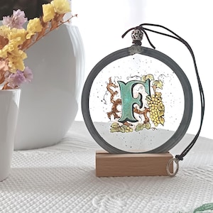May include: A round, decorative glass ornament with the letter "F" in teal, surrounded by grape vines and grapes. The ornament has a dark gray metal frame and hangs from a black cord. It rests on a small wooden stand.