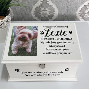 May include: A white wooden pet memorial box featuring a photo of a small dog named Lexie. The box is inscribed with "Treasured Memories Of Lexie", dates, and a touching message. Paw prints decorate the box.