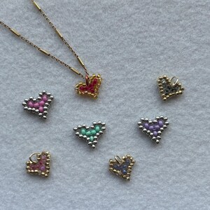 Puede incluir: Un collar dorado con un colgante de coraz&oacute;n de cuentas rojas y doradas. Varios dijes de coraz&oacute;n de cuentas en rosa, verde, morado y dorado est&aacute;n dispersos. Las cuentas son peque&ntilde;as y crean un efecto texturizado.