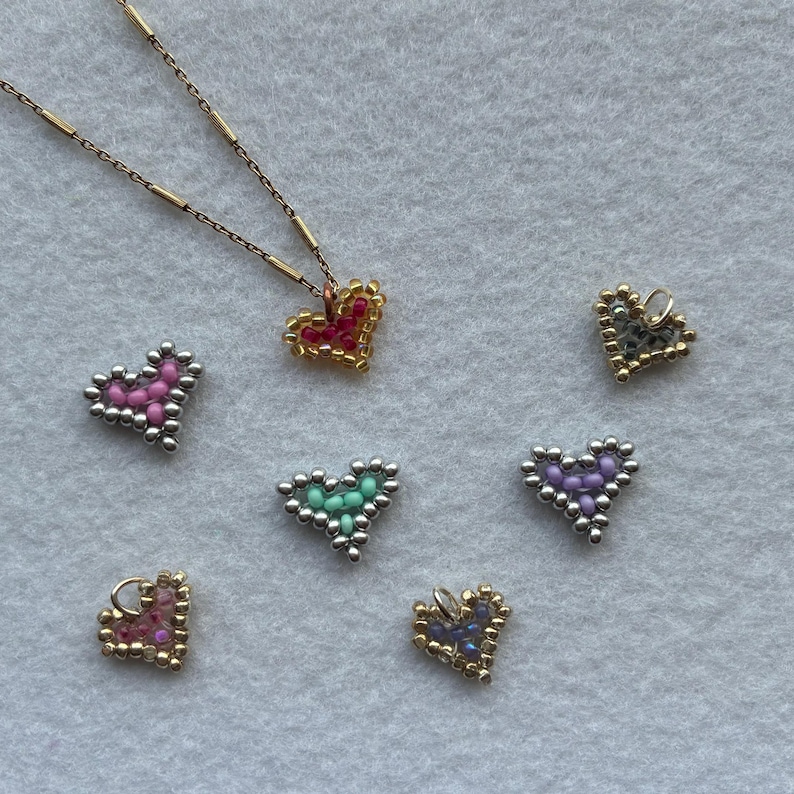 Puede incluir: Un collar dorado con un colgante de coraz&oacute;n de cuentas rojas y doradas. Varios dijes de coraz&oacute;n de cuentas en rosa, verde, morado y dorado est&aacute;n dispersos. Las cuentas son peque&ntilde;as y crean un efecto texturizado.