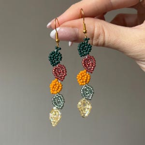 Pu&ograve; includere: Un paio di orecchini di perline con ganci color oro. Ogni orecchino presenta una serie di forme di perline colorate: verde scuro, rosso, arancione, argento e giallo. Gli orecchini sono fatti a mano e hanno un design artistico unico.