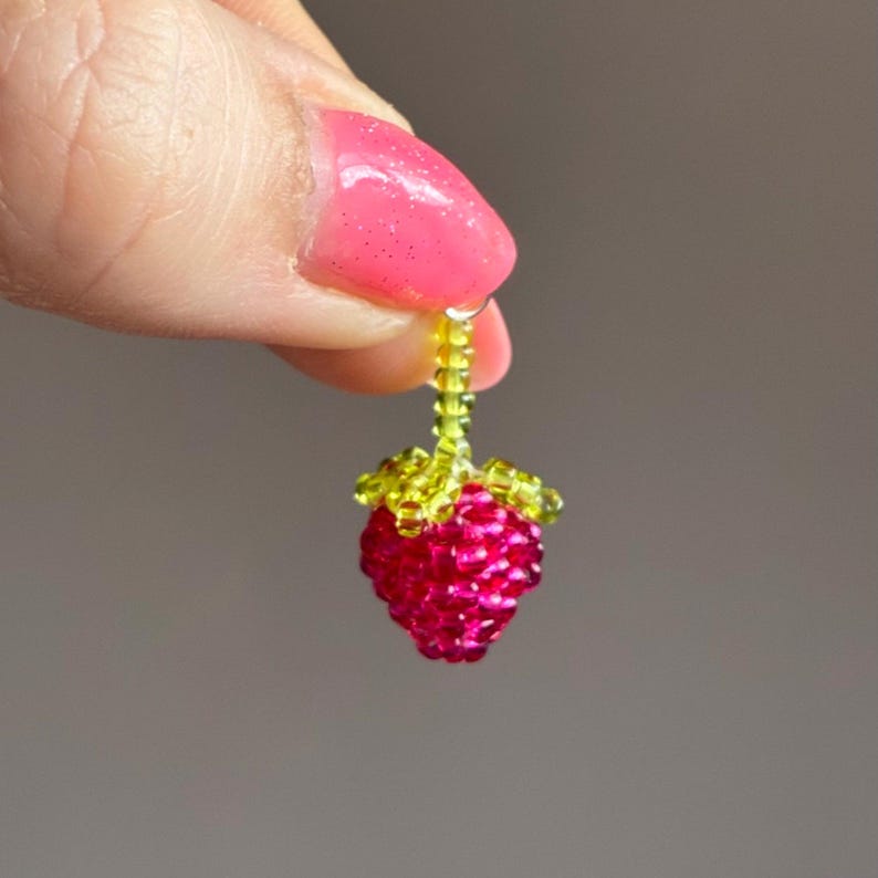 May include: A small, three-dimensional raspberry charm crafted from tiny, vibrant pink glass beads. The fruit features a cluster of bright lime green beads at the top, forming the stem and leaves, creating a detailed, textured berry shape.