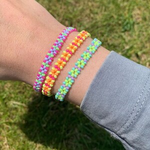 May include: Three colorful beaded bracelets stacked on a wrist. The bracelets are made with small, bright pink, orange, yellow, green, and blue beads.