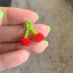 May include: A pair of cherry earrings. Each cherry is made of small red beads, with green beaded stems and a silver metal ring. The earrings are a playful accessory.