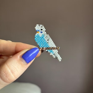Op de afbeelding: Een kralen parkiet figuur in blauw, wit en grijs, zittend op een bruine kralen tak. De vogel heeft een gele snavel en zwarte ogen. Het beeldje wordt vastgehouden door een hand met blauwe nagellak.
