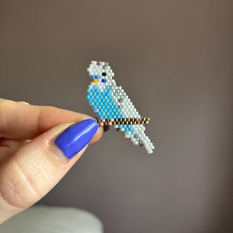 Op de afbeelding: Een kralen parkiet figuur in blauw, wit en grijs, zittend op een bruine kralen tak. De vogel heeft een gele snavel en zwarte ogen. Het beeldje wordt vastgehouden door een hand met blauwe nagellak.