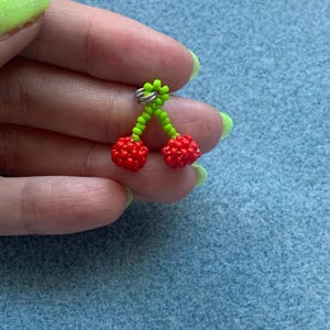 May include: A pair of cherry earrings. The earrings feature two red beaded cherries with green beaded stems and a silver-colored earring hook. The earrings are held against a blue background.