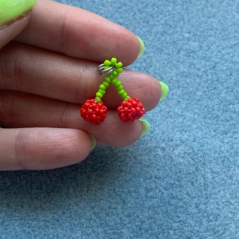 May include: A pair of cherry earrings. The earrings feature two red beaded cherries with green beaded stems and a silver-colored earring hook. The earrings are held against a blue background.