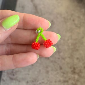 May include: A beaded cherry earring with red cherries and green stems. The cherries are made of small red beads, and the stems are made of green beads. The earring has a silver-colored metal loop.
