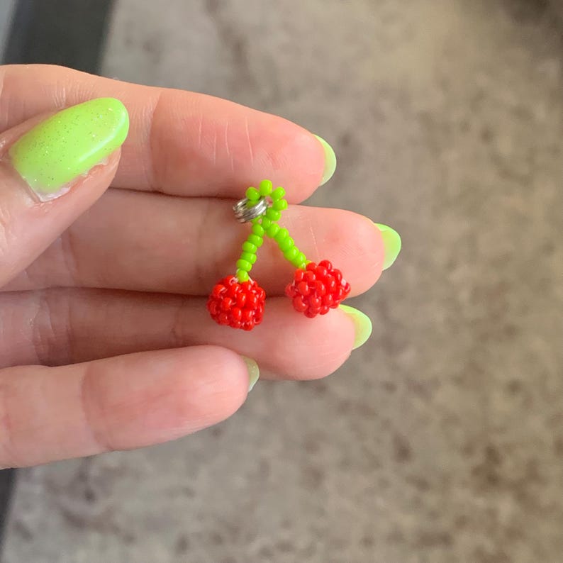 May include: A beaded cherry earring with red cherries and green stems. The cherries are made of small red beads, and the stems are made of green beads. The earring has a silver-colored metal loop.