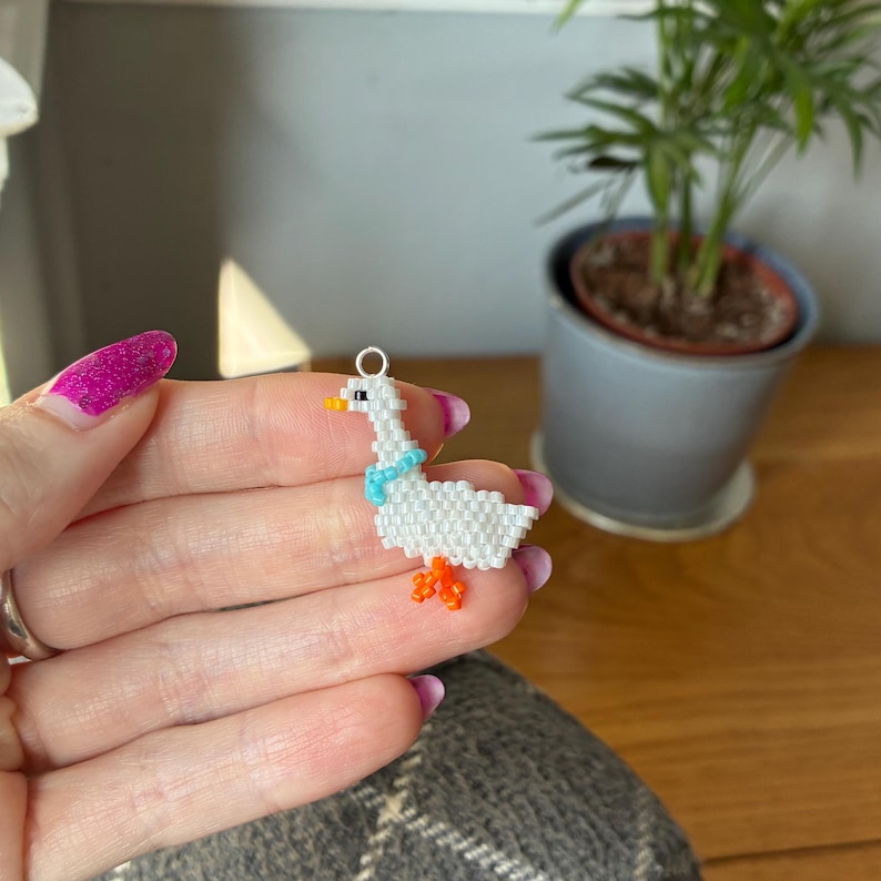 Puede incluir: Un charm de ganso hecho a mano con cuentas blancas, con una bufanda azul claro y patas naranjas. El ganso tiene un pico amarillo y un lazo plateado para colgar. Ideal para la creaci&oacute;n de joyas o como un accesorio &uacute;nico.