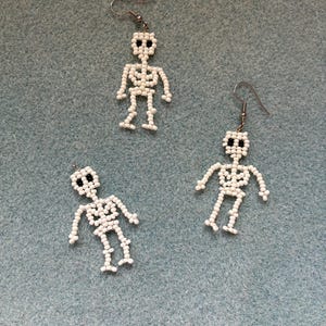 May include: Three pairs of white beaded skeleton earrings with black bead eyes and silver-tone ear wires. The earrings are arranged on a light blue textured surface. The skeleton earrings are a fun accessory for Halloween or everyday wear.