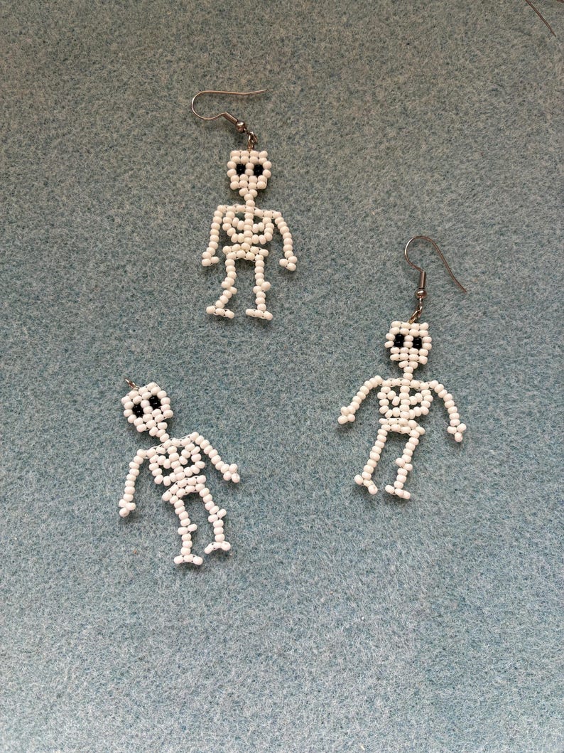 May include: Three pairs of white beaded skeleton earrings with black bead eyes and silver-tone ear wires. The earrings are arranged on a light blue textured surface. The skeleton earrings are a fun accessory for Halloween or everyday wear.