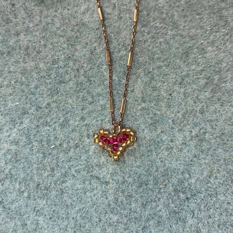 Puede incluir: Un collar dorado con un colgante en forma de coraz&oacute;n. El colgante est&aacute; hecho de peque&ntilde;as cuentas doradas que delinean un coraz&oacute;n lleno de cuentas rojas. La cadena del collar tiene un dise&ntilde;o delicado con peque&ntilde;as barras doradas.