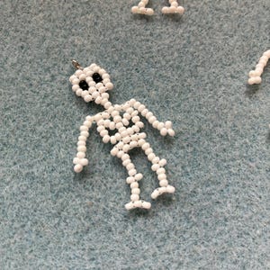 May include: White beaded skeleton charm with black bead eyes. The skeleton is made of small, round white beads strung together to form the shape of a human skeleton. The charm has a small silver loop at the top for hanging. Several other skeleton charms are visible.