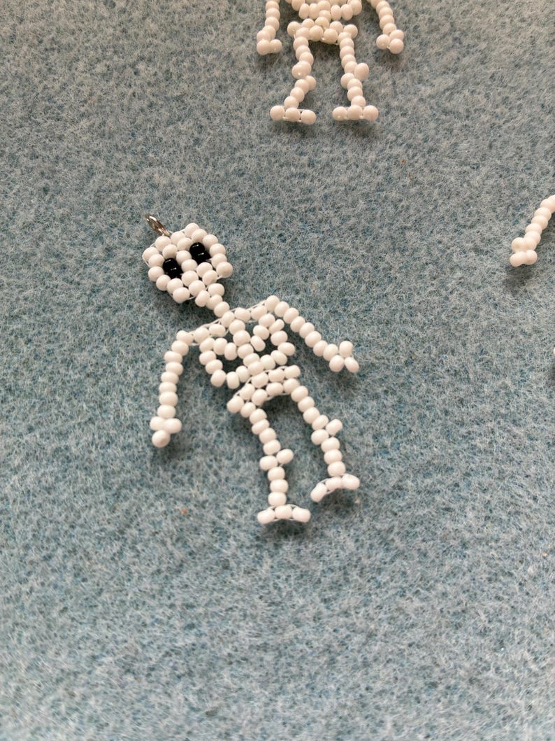 May include: White beaded skeleton charm with black bead eyes. The skeleton is made of small, round white beads strung together to form the shape of a human skeleton. The charm has a small silver loop at the top for hanging. Several other skeleton charms are visible.