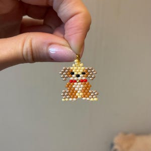May include: A beaded dog charm in shades of brown, tan, and cream, with a red bow tie. The charm is made of small beads and has a gold-colored loop at the top for hanging. The dog's features are detailed with black beads.
