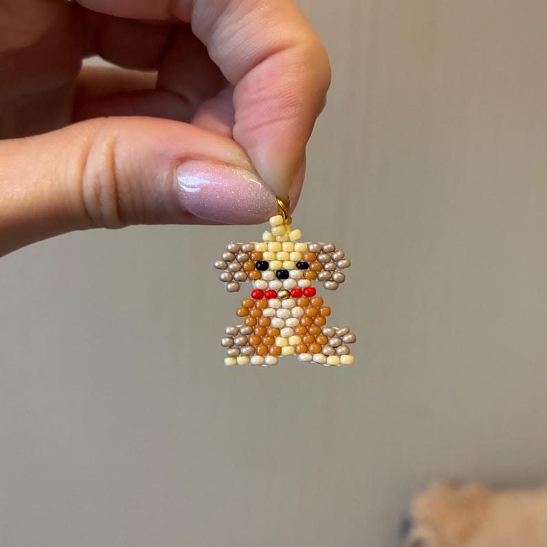 May include: A beaded dog charm in shades of brown, tan, and cream, with a red bow tie. The charm is made of small beads and has a gold-colored loop at the top for hanging. The dog's features are detailed with black beads.