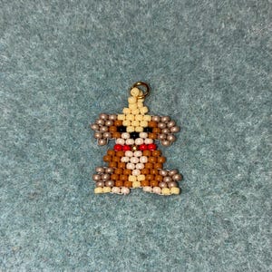 May include: A beaded dog charm with a gold-colored loop for hanging. The dog is made of tan, brown, black, white, and red beads. The charm has a red collar and tan ears. The background is a teal-colored fabric.
