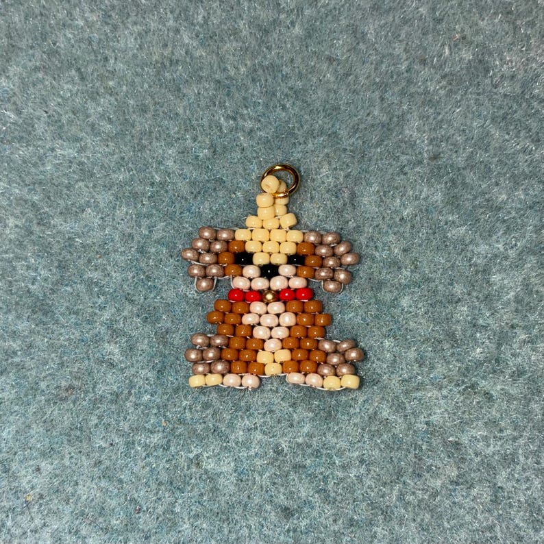 May include: A beaded dog charm with a gold-colored loop for hanging. The dog is made of tan, brown, black, white, and red beads. The charm has a red collar and tan ears. The background is a teal-colored fabric.
