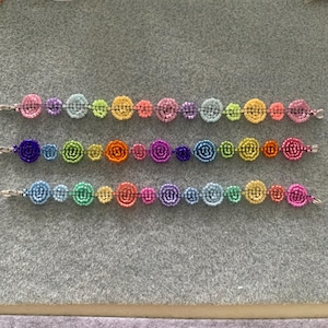Pode incluir: Três pulseiras de macramé coloridas com uma série de círculos de contas em tons de rosa, azul, amarelo, verde e roxo. Cada pulseira tem um fecho prateado. As pulseiras são exibidas em uma superfície cinza.