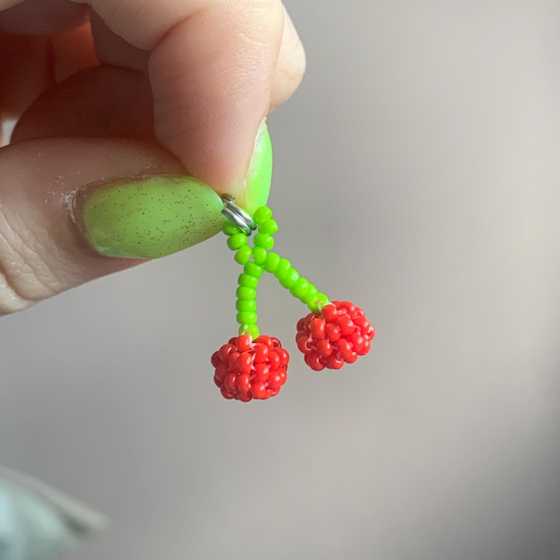 May include: A pair of beaded cherry earrings. The cherries are made of small red beads, and the stems are made of green beads. The earrings are attached to a silver-colored ring.
