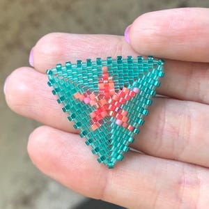 May include: A handmade, beaded triangle pendant featuring a turquoise background with a coral starfish design in the center. The pendant is made with small, colorful beads and is held in a hand.