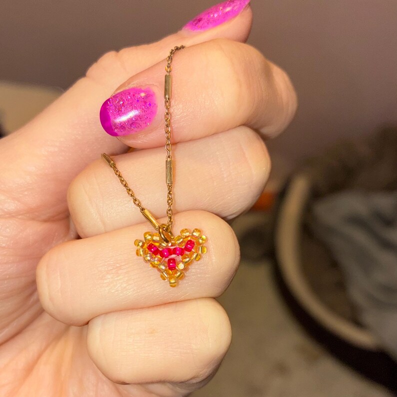 Puede incluir: Un collar dorado con un colgante en forma de coraz&oacute;n. El colgante est&aacute; hecho de peque&ntilde;as cuentas doradas y rojas. El collar se sostiene en una mano con esmalte de u&ntilde;as rosa brillante.
