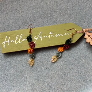 Pu&ograve; includere: Un paio di orecchini pendenti con perline dai colori autunnali. Gli orecchini presentano perline verdi, rosse, arancioni e dorate. Sono esposti su un'etichetta di legno verde con la scritta "Hello Autumn".
