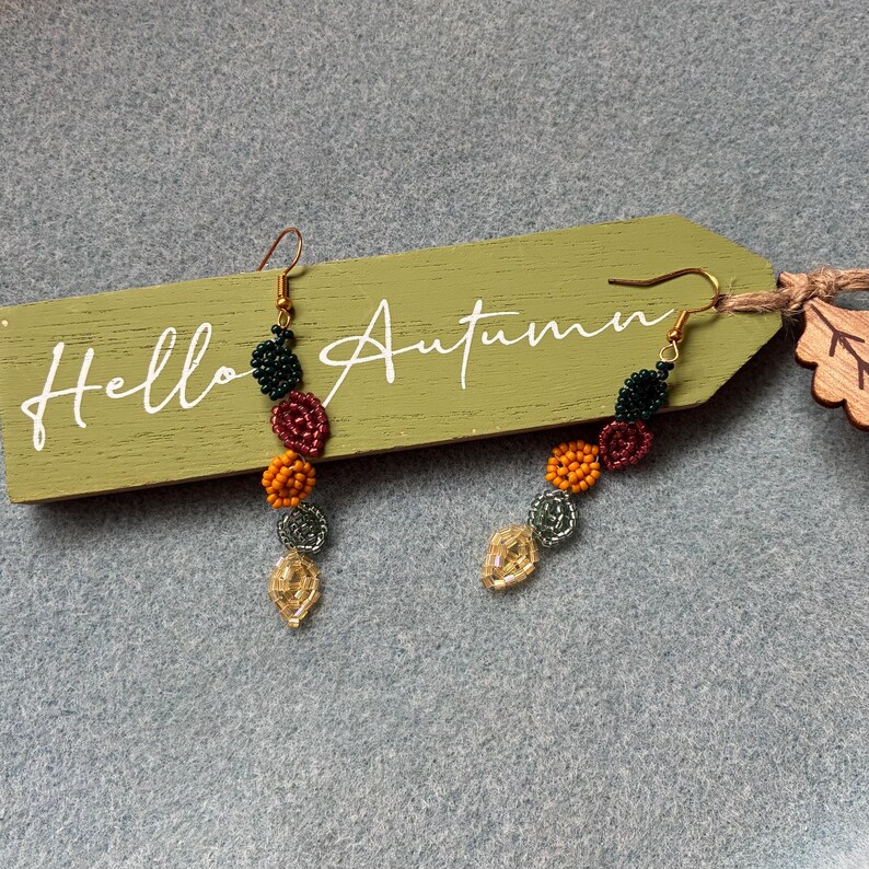 Pu&ograve; includere: Un paio di orecchini pendenti con perline dai colori autunnali. Gli orecchini presentano perline verdi, rosse, arancioni e dorate. Sono esposti su un'etichetta di legno verde con la scritta "Hello Autumn".