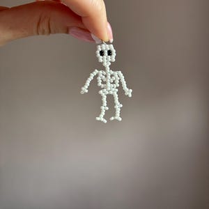 May include: A small, white beaded skeleton figure with black bead eyes. The skeleton is held up by a small loop at the top, suggesting it could be a charm or pendant. The figure is made of small, round beads.