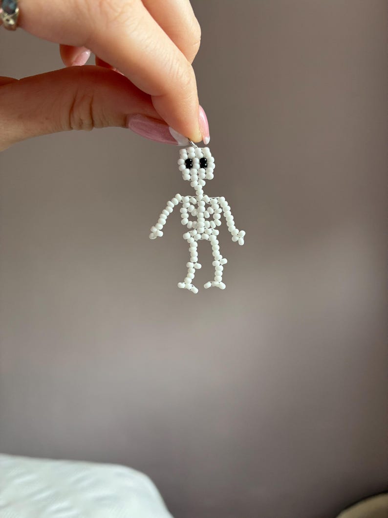 May include: A small, white beaded skeleton figure with black bead eyes. The skeleton is held up by a small loop at the top, suggesting it could be a charm or pendant. The figure is made of small, round beads.