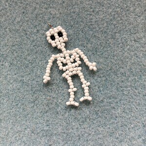 May include: A handmade skeleton charm made of white beads with black bead eyes. The skeleton is strung together with a silver-colored metal loop at the top for hanging. The charm is a Halloween or gothic-themed accessory.