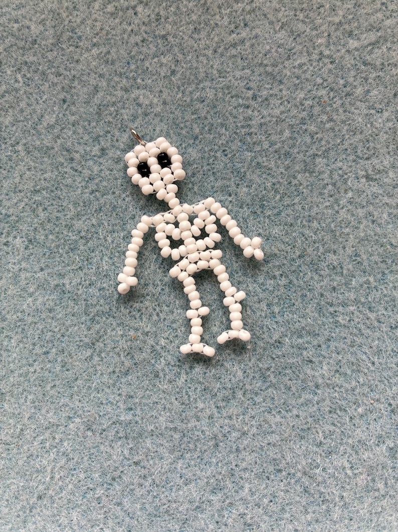 May include: A handmade skeleton charm made of white beads with black bead eyes. The skeleton is strung together with a silver-colored metal loop at the top for hanging. The charm is a Halloween or gothic-themed accessory.