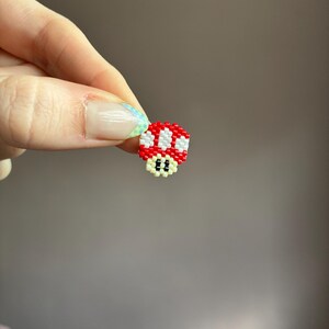 Pode incluir: Um pequeno pingente de cogumelo pixelado feito de contas vermelhas, brancas e pretas. O pingente &eacute; uma representa&ccedil;&atilde;o em miniatura de um personagem de videogame. O cogumelo tem um chap&eacute;u vermelho com pontos brancos e uma base preta com dois olhos pequenos.