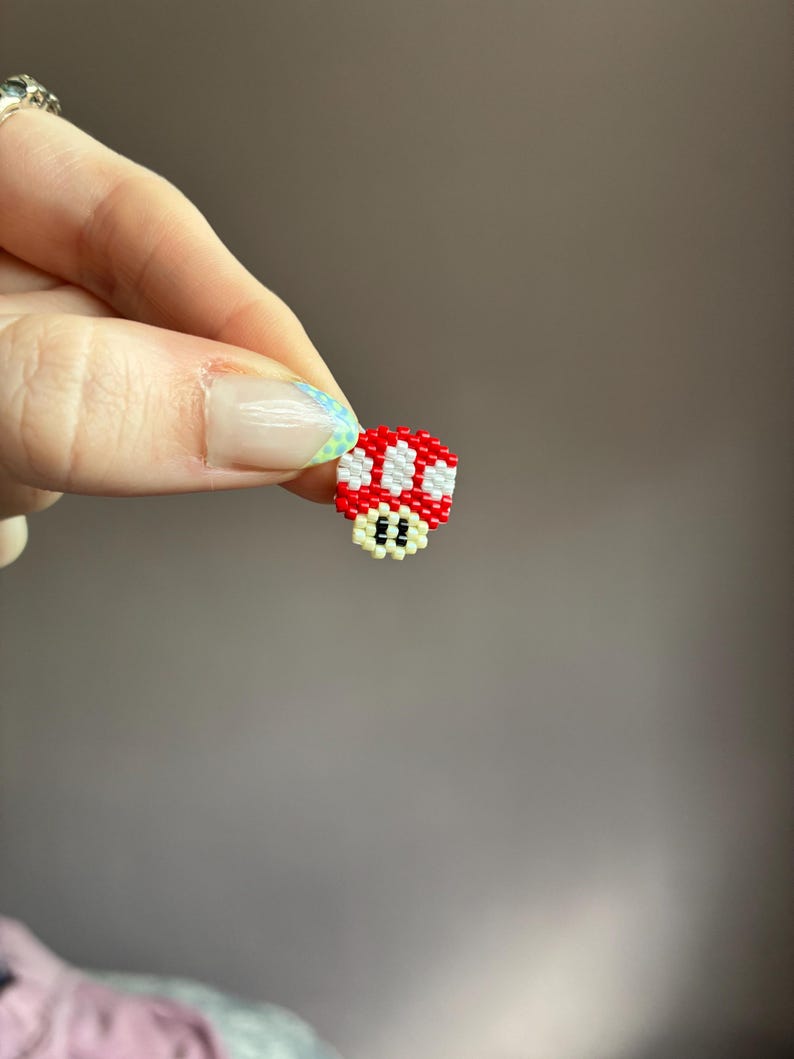 Pode incluir: Um pequeno pingente de cogumelo pixelado feito de contas vermelhas, brancas e pretas. O pingente &eacute; uma representa&ccedil;&atilde;o em miniatura de um personagem de videogame. O cogumelo tem um chap&eacute;u vermelho com pontos brancos e uma base preta com dois olhos pequenos.