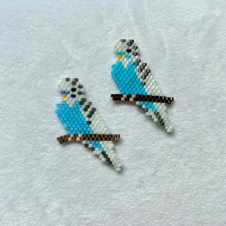Op de afbeelding: Twee kralen parkiet ornamenten. Elke vogel is turquoise, wit en grijs, zittend op een bruine, met kralen bezette tak. De parkieten hebben gele en zwarte accenten. Handgemaakte ornamenten voor interieurdecoratie.