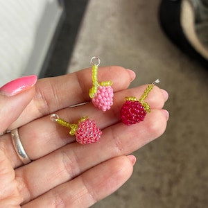 May include: Three small charms shaped like raspberries, crafted from tiny glass beads. One charm is light pink, one is bright red, and one is a darker reddish-pink. Each charm features a green beaded stem and a small silver metal jump ring.
