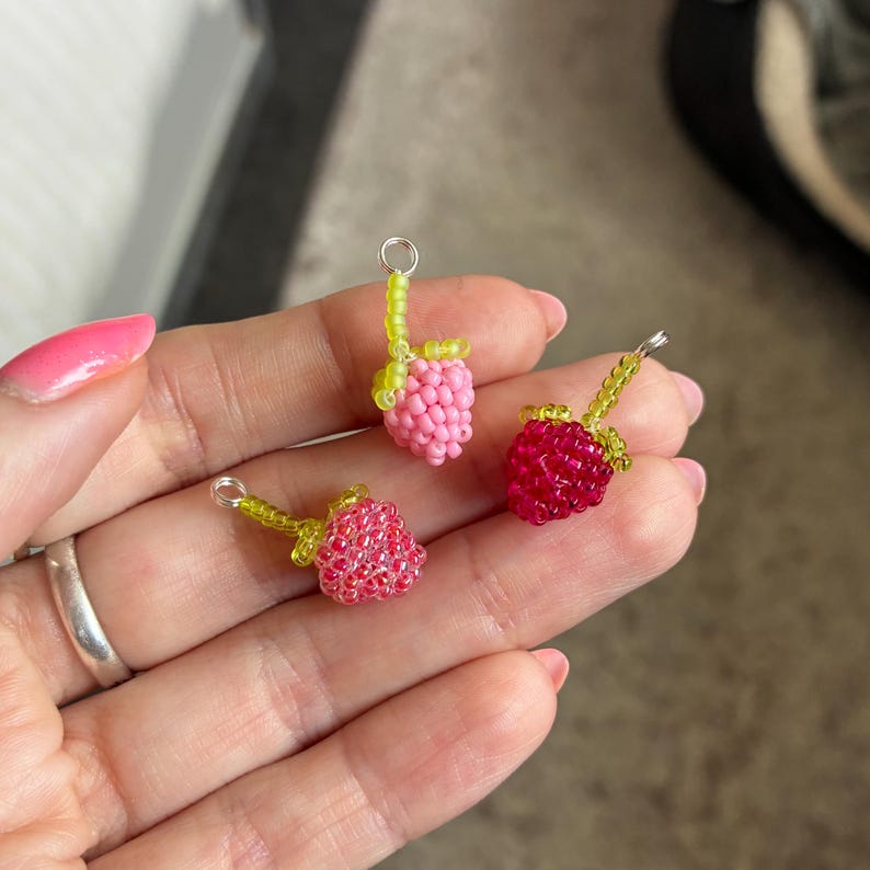 May include: Three small charms shaped like raspberries, crafted from tiny glass beads. One charm is light pink, one is bright red, and one is a darker reddish-pink. Each charm features a green beaded stem and a small silver metal jump ring.