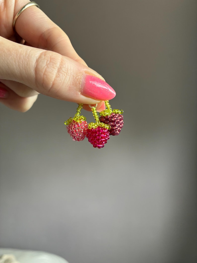 May include: A cluster of three small berry charms crafted from tiny glass beads in shades of light pink, vibrant magenta, and deep maroon. Each berry features a small green beaded stem and leaf detail, connected together by a small metal jump ring.