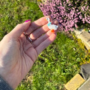 Puede incluir: Un charm de ganso hecho a mano con cuentas. El charm tiene un cuerpo blanco, una bufanda azul y patas naranjas. Tiene un lazo plateado para colgar. El fondo muestra hierba verde y flores moradas.