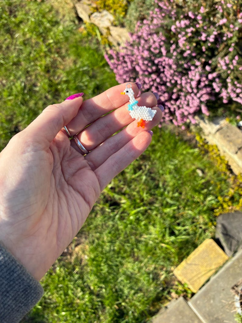 Puede incluir: Un charm de ganso hecho a mano con cuentas. El charm tiene un cuerpo blanco, una bufanda azul y patas naranjas. Tiene un lazo plateado para colgar. El fondo muestra hierba verde y flores moradas.