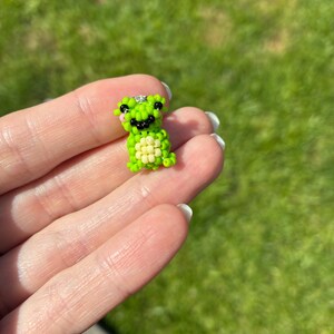Puede incluir: Peque&ntilde;a figura de rana hecha con cuentas en tonos verdes y amarillos. La rana tiene ojos negros y una boca sonriente. La figura se sostiene en una mano sobre un fondo verde borroso.