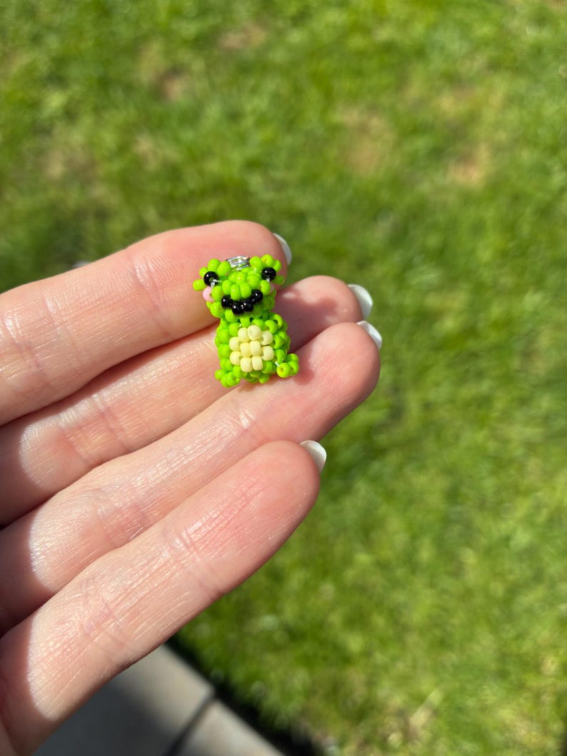 Puede incluir: Peque&ntilde;a figura de rana hecha con cuentas en tonos verdes y amarillos. La rana tiene ojos negros y una boca sonriente. La figura se sostiene en una mano sobre un fondo verde borroso.