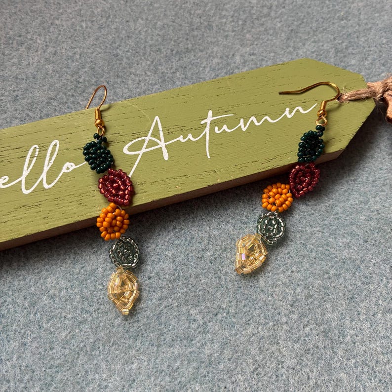 Pu&ograve; includere: Un paio di orecchini di perline a tema autunnale. Gli orecchini presentano una serie di perline colorate nelle tonalit&agrave; del verde, del rosso, dell'arancione, dell'argento e dell'oro. Gli orecchini sono appesi a ganci color oro. Lo sfondo include un'etichetta di legno che dice "Autumn".