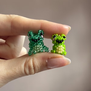 Puede incluir: Dos figuras de rana hechas con cuentas. Una es verde transl&uacute;cido, la otra es verde lima con vientre color crema. Ambas tienen ojos negros y mejillas rosas. Las ranas son peque&ntilde;as, probablemente de menos de 2,5 cm de altura, y est&aacute;n hechas a mano.