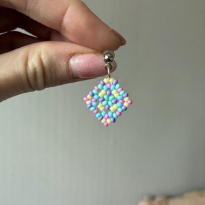 May include: A single beaded earring with a silver post. The earring features a diamond-shaped design made of small, colorful beads in pastel shades of pink, yellow, blue, and purple. The earring is held by a hand.